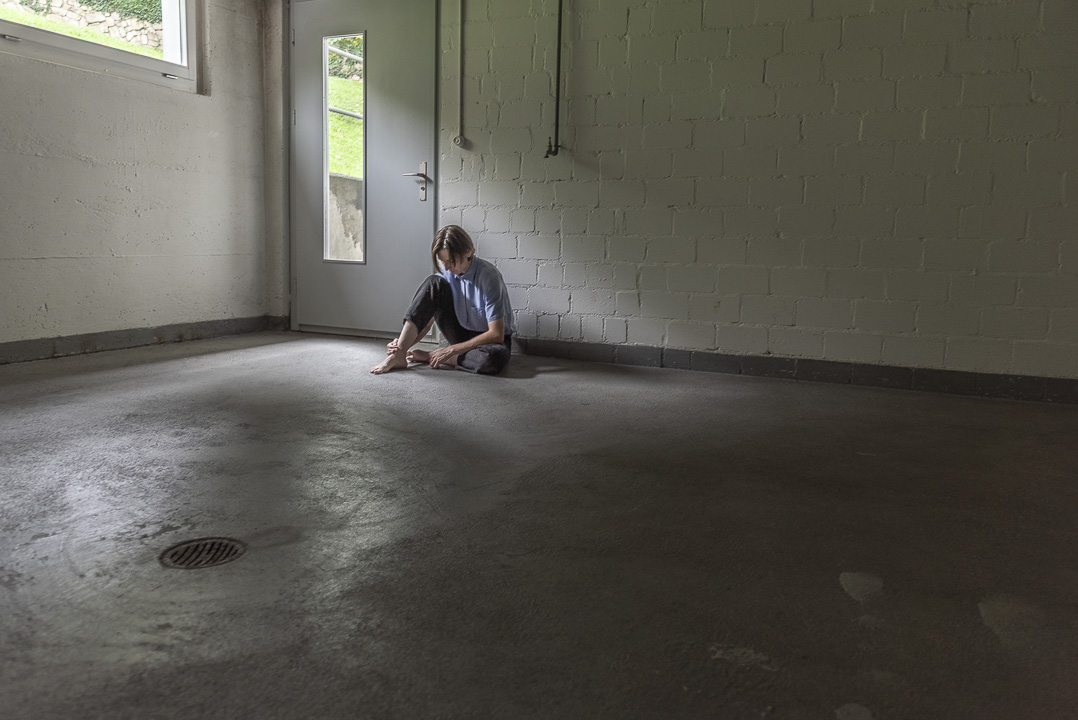 Junger Mann sitzt in einem dunklen Raum hinten an der Wand am Boden, das rechte Bein aufgestellt und senkt den Kopf. Jacqueline Morgenroth Fotografie